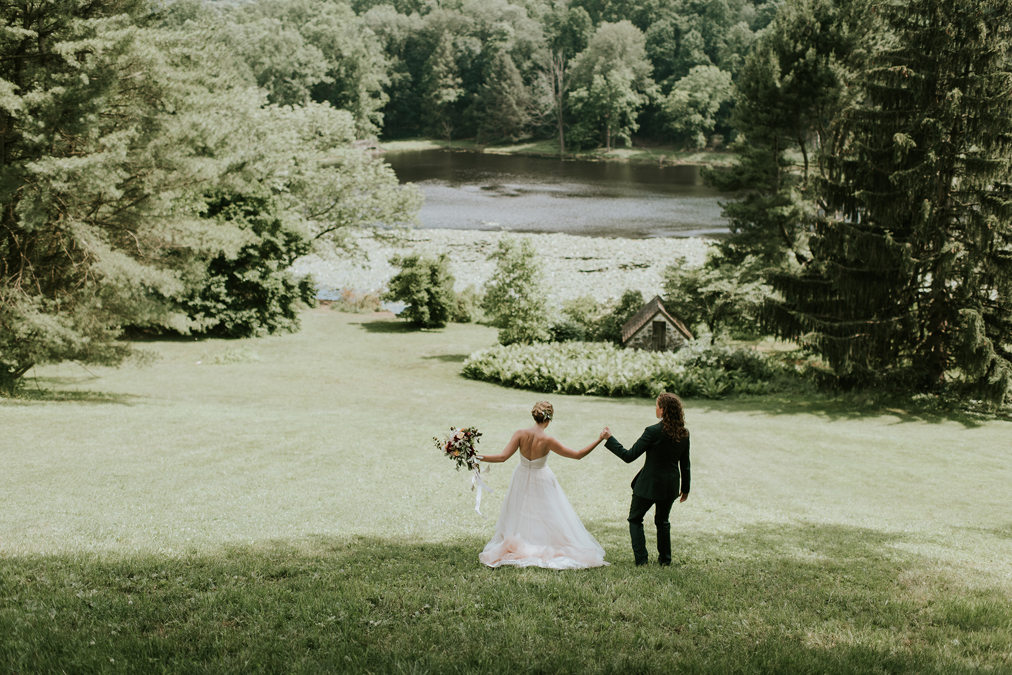 A newlywed couple holding hands across a wide lawn with trees and a pond behind them