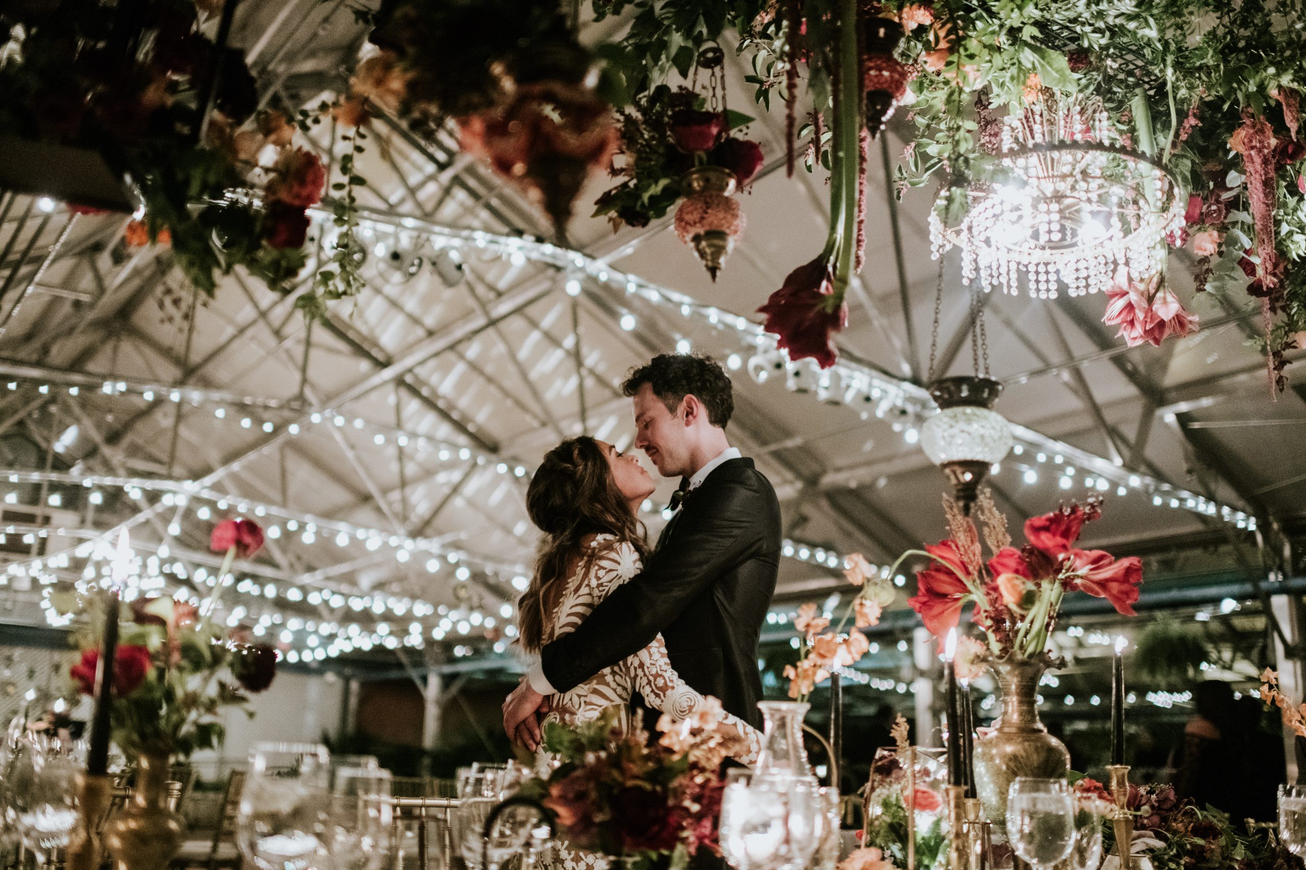 A newlywed couple embracing beneath suspended flowers and chandeliers at the Horticulture Center