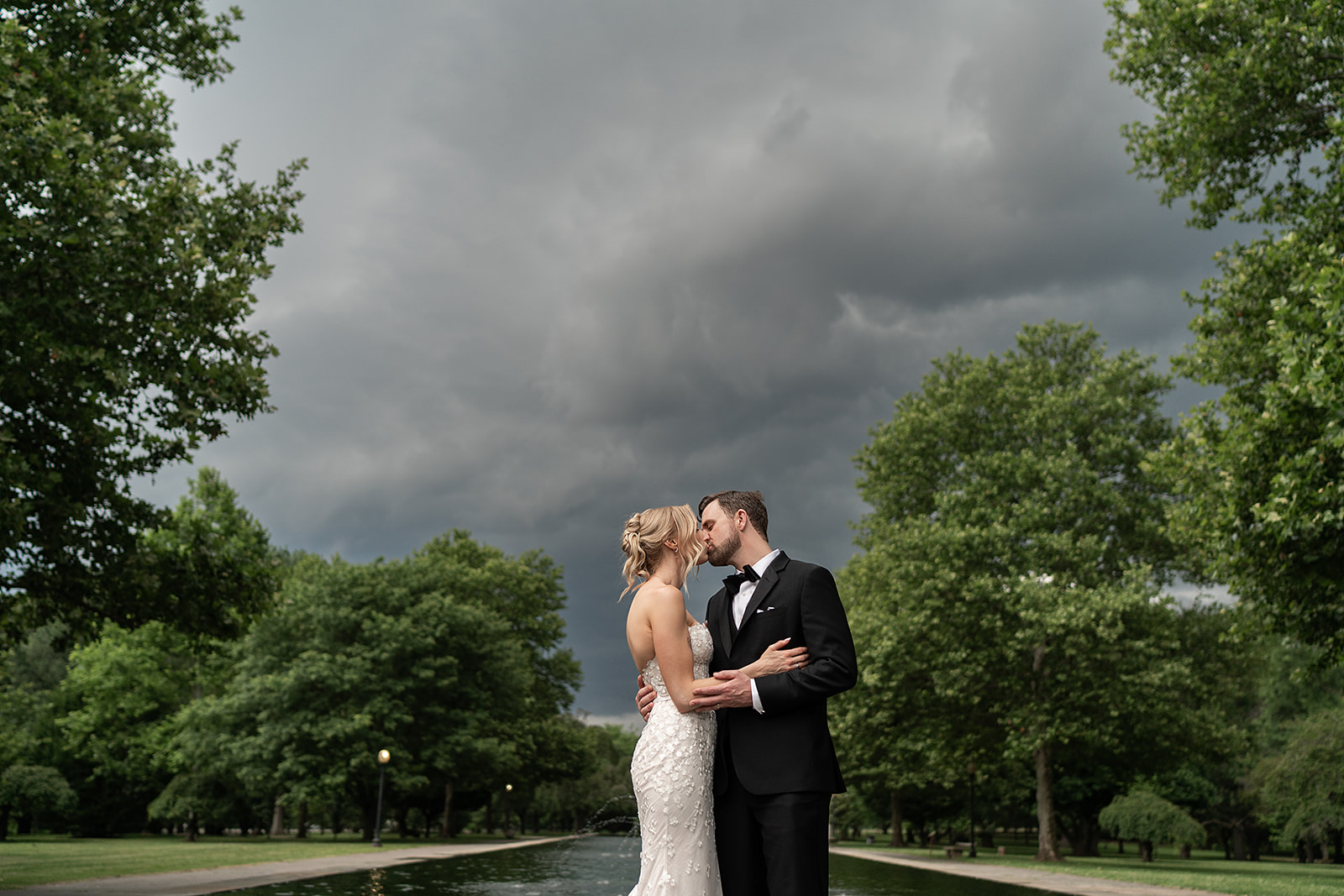 A newlywed couple kissing beneath storm clouds beside the Horticulture Center reflecting pool